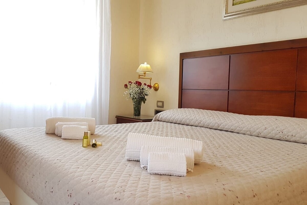 A neatly made bed with white linens, a bedside table with a lamp and a vase of flowers, and a wooden headboard.