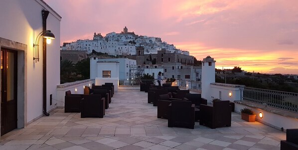 Terraza en la azotea con mobiliario exterior y vistas a un pueblo blanco en la cima al atardecer.