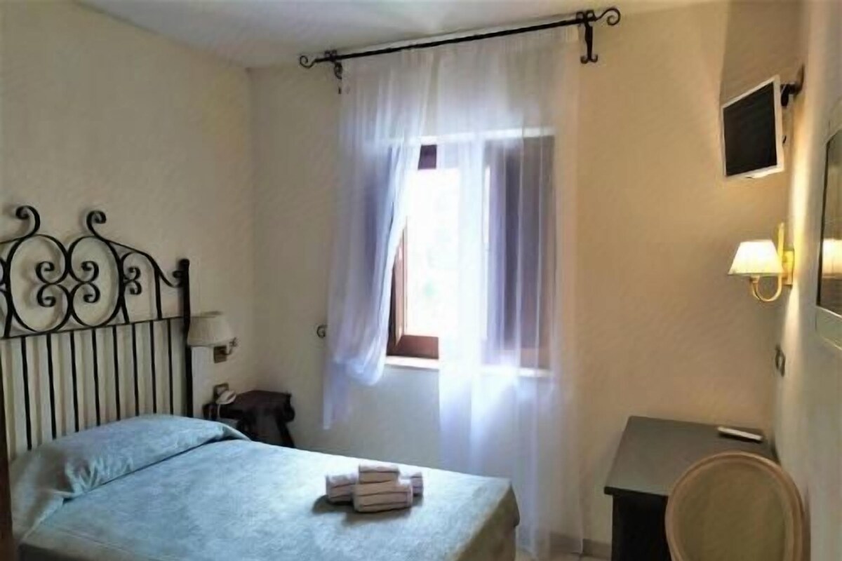 Un dormitorio con cama, una silla, una mesita de noche, una lámpara y una ventana con cortinas.
