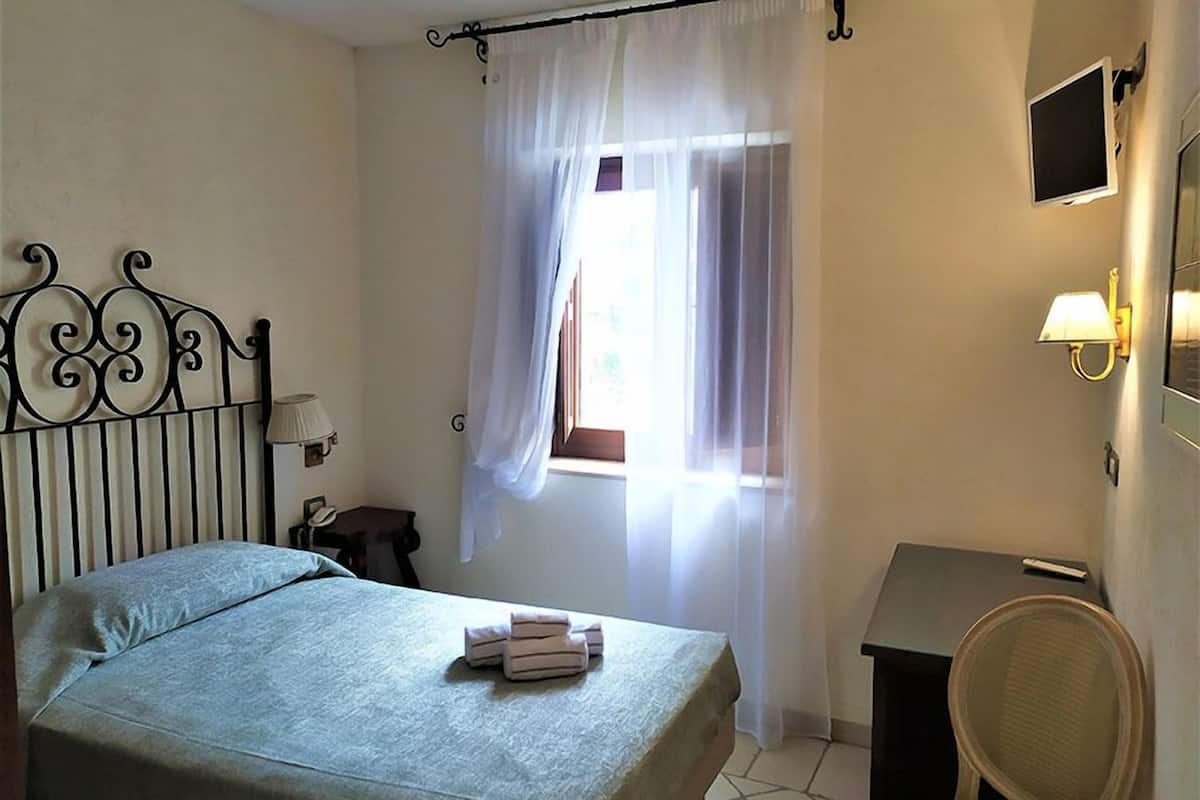 A bedroom with a bed, a bedside table, a chair, a desk, a window with curtains, and a wall-mounted TV.