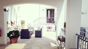 Un patio luminoso y soleado con mobiliario exterior, plantas en macetas y una escalera que lleva a un nivel superior.