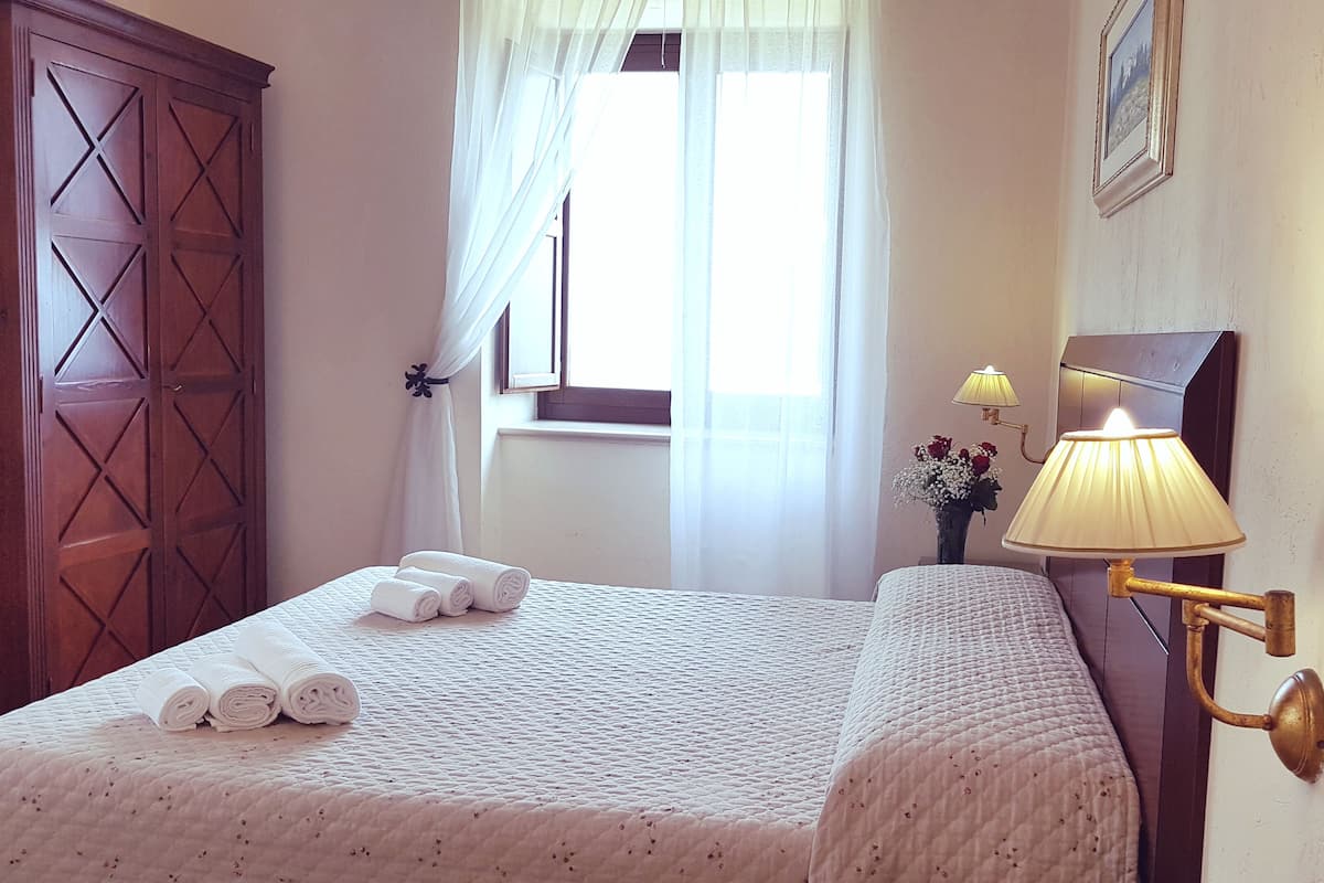 A neatly made bed with rolled towels, a window with sheer curtains, a wall-mounted lamp, and a wooden wardrobe.