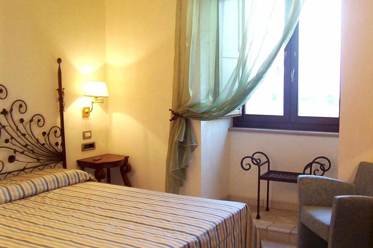 A hotel room with a bed, a nightstand, a chair, a lamp, and a window with curtains.