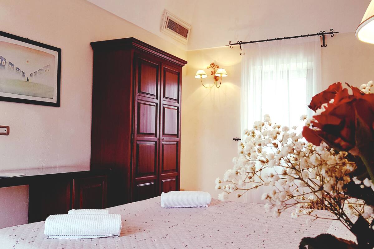 A hotel room with a bed, a wardrobe, a wall-mounted lamp, a window with curtains, and a framed picture on the wall.