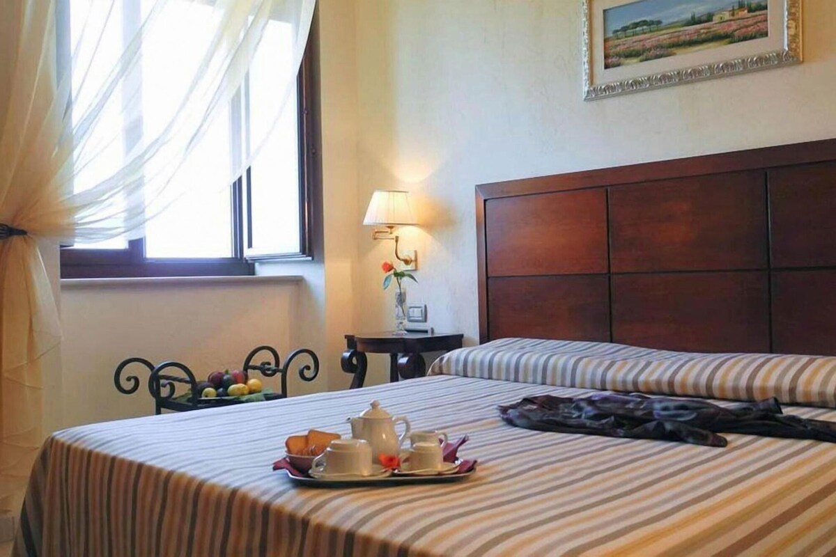 A hotel room with two beds, a bedside table with a lamp, a framed picture on the wall, and a window with sheer curtains.