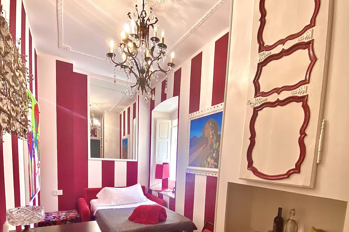 A hotel room with a bed, a desk, a chair, a chandelier, and a painting on the wall.