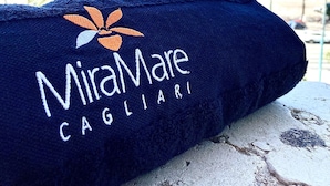 A dark blue towel with the text "MiraMare Cagliari" and a logo, placed on a stone surface overlooking a marina with boats and palm trees.