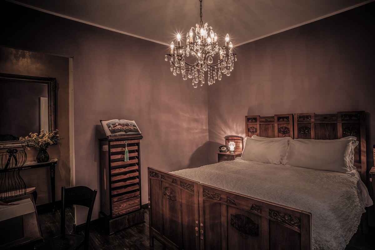 Une chambre à coucher avec un lit en bois, un lustre, une table de chevet avec une lampe et un livre posé sur une petite table d’appoint.