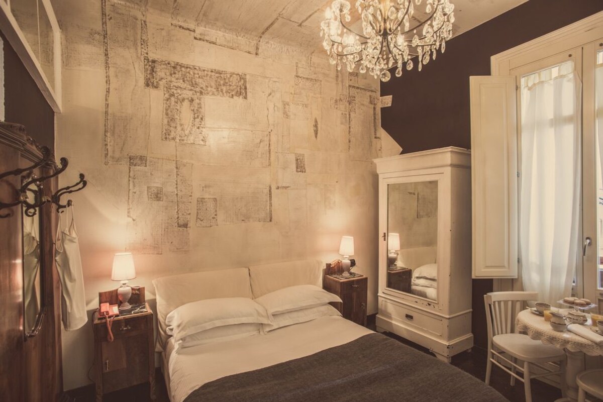 Une chambre d’hôtel avec un lit, des tables de chevet, un miroir, un lustre et une fenêtre avec des rideaux.