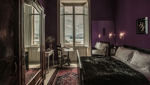A bedroom with a large bed, a window with a view, a chair, a small table, and a dresser.