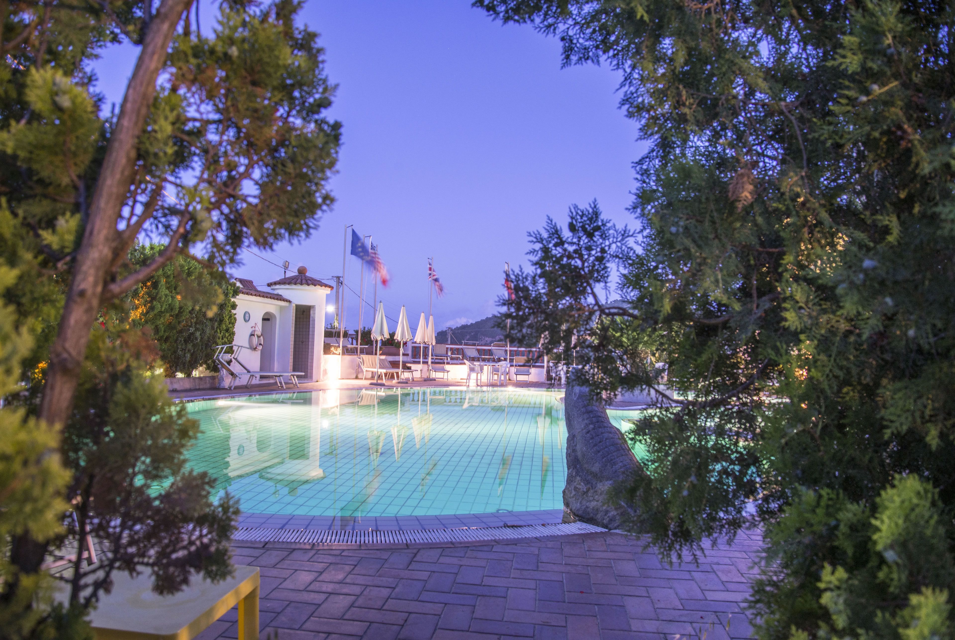Ein Swimmingpool mit Liegestühlen und Sonnenschirmen, umgeben von Bäumen und einem Gebäude mit Flaggen.