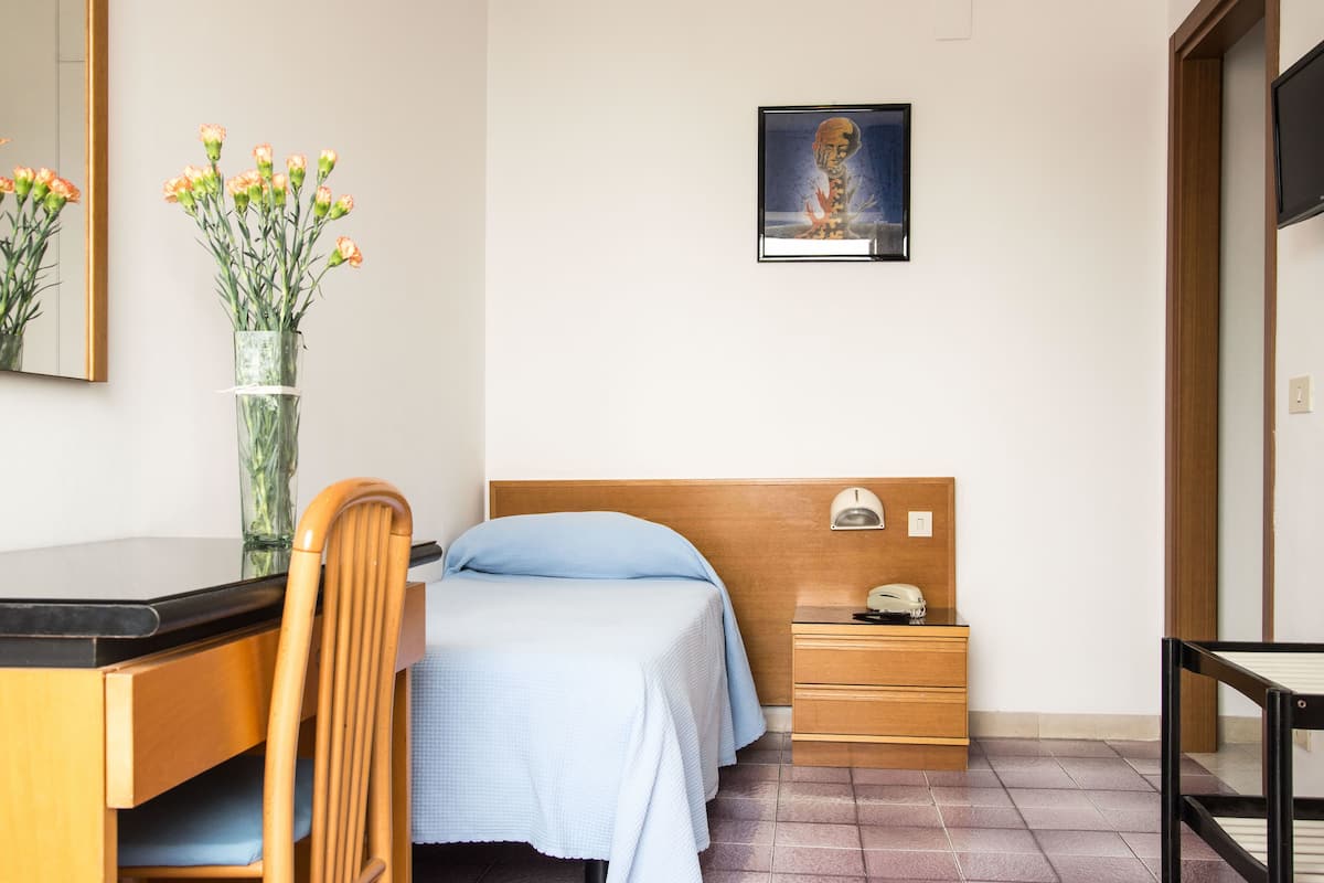 A bedroom with a bed, a nightstand, a desk, a chair, a vase with flowers, and a framed picture on the wall.