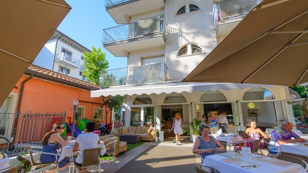 An outdoor cafe with tables and chairs, a building with balconies, and a clear sky. - Falco (Riccione)