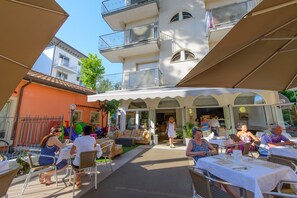 An outdoor cafe with tables and chairs, a building with balconies, and a clear sky. - Falco (Riccione)
