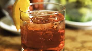 Un verre d’une boisson ambrée, agrémentée de glaçons et d’une tranche d’orange, posé sur une table recouverte d’une nappe blanche en dentelle.