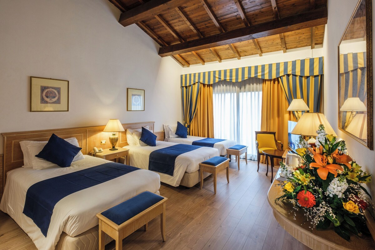 Habitación de hotel con dos camas, suelo de madera, techo de madera, un escritorio con silla, un espejo y un arreglo floral.