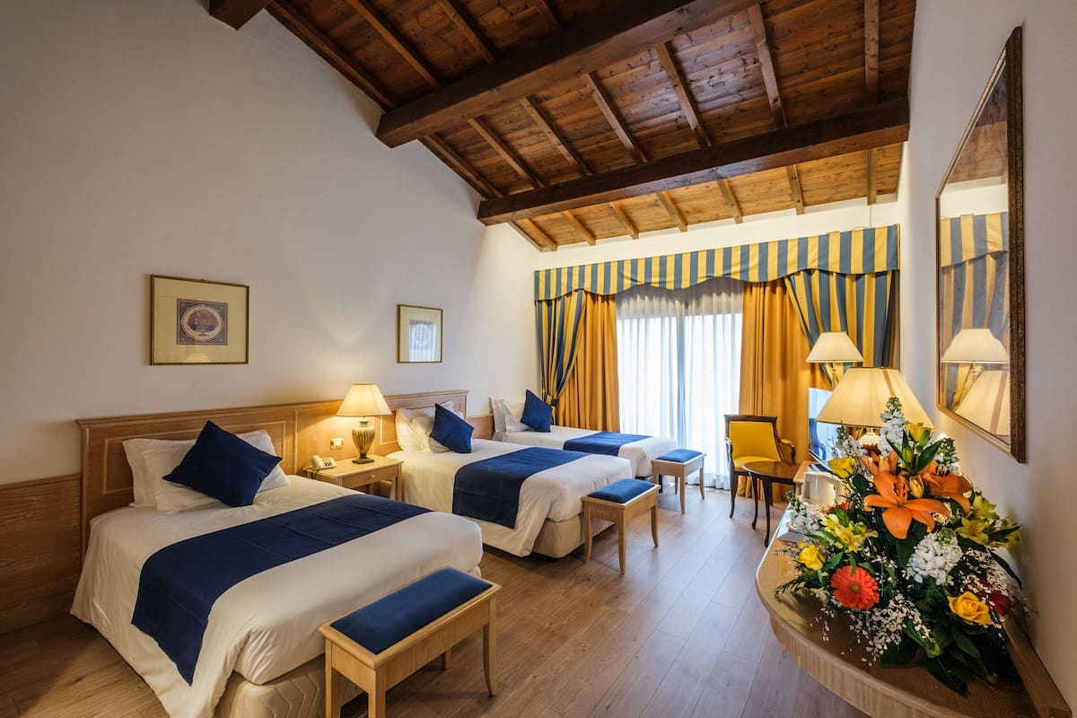 Habitación de hotel con tres camas, cabeceras de madera y techo de madera. Hay mesitas de noche con lámparas, una pequeña mesa con un arreglo floral y una silla.
