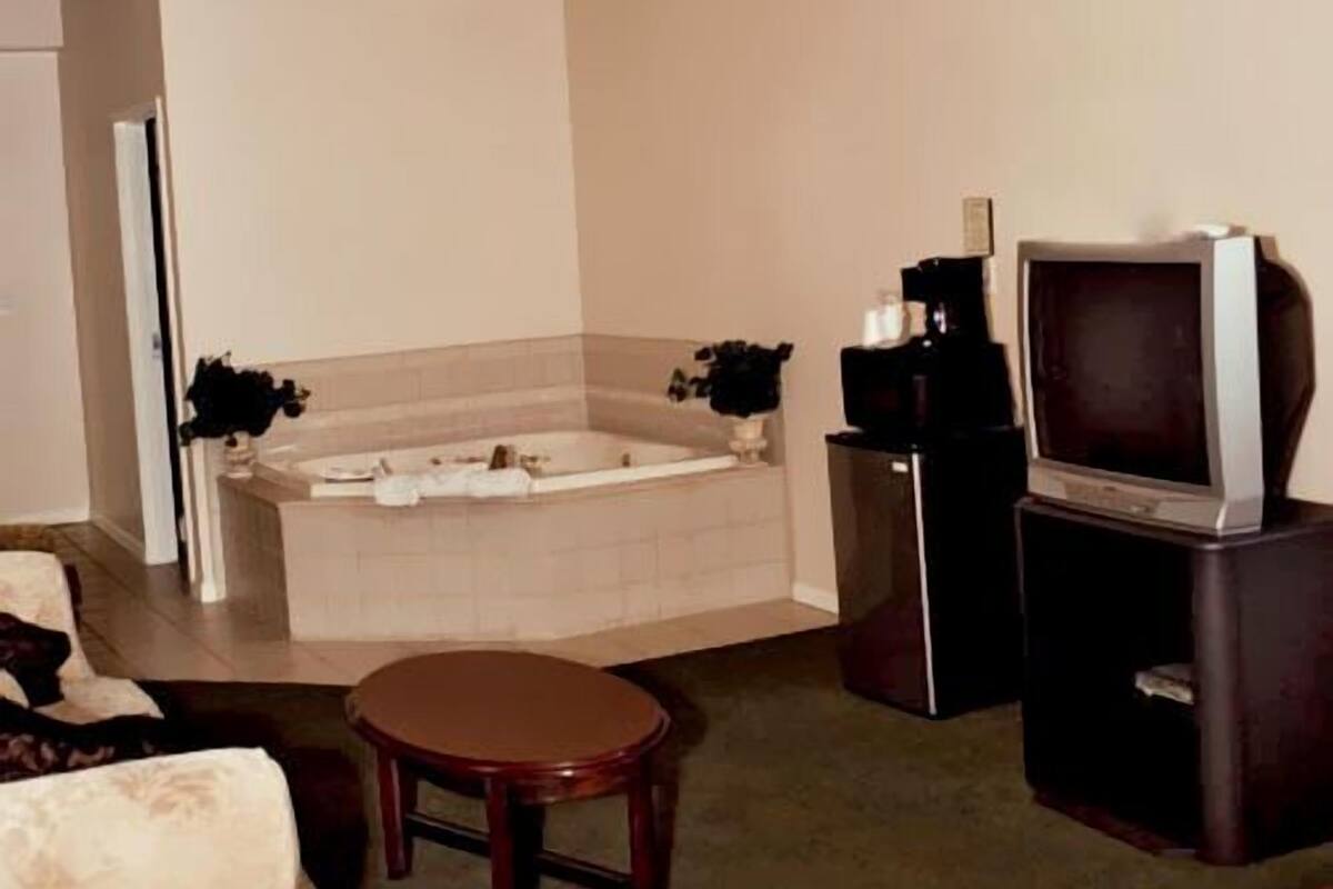 A hotel room with a bed, a small round table, a TV on a stand, a bathtub, and a desk with a chair.