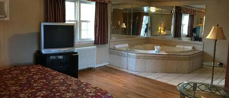 Una habitación de hotel con cama, televisión, espejo grande, bañera y mesa de vidrio.