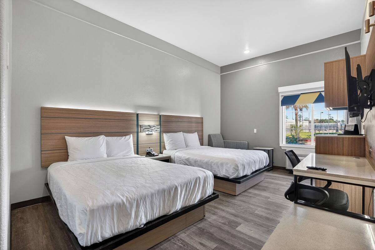 A modern hotel room with two beds, a desk, and a view of the outdoors.