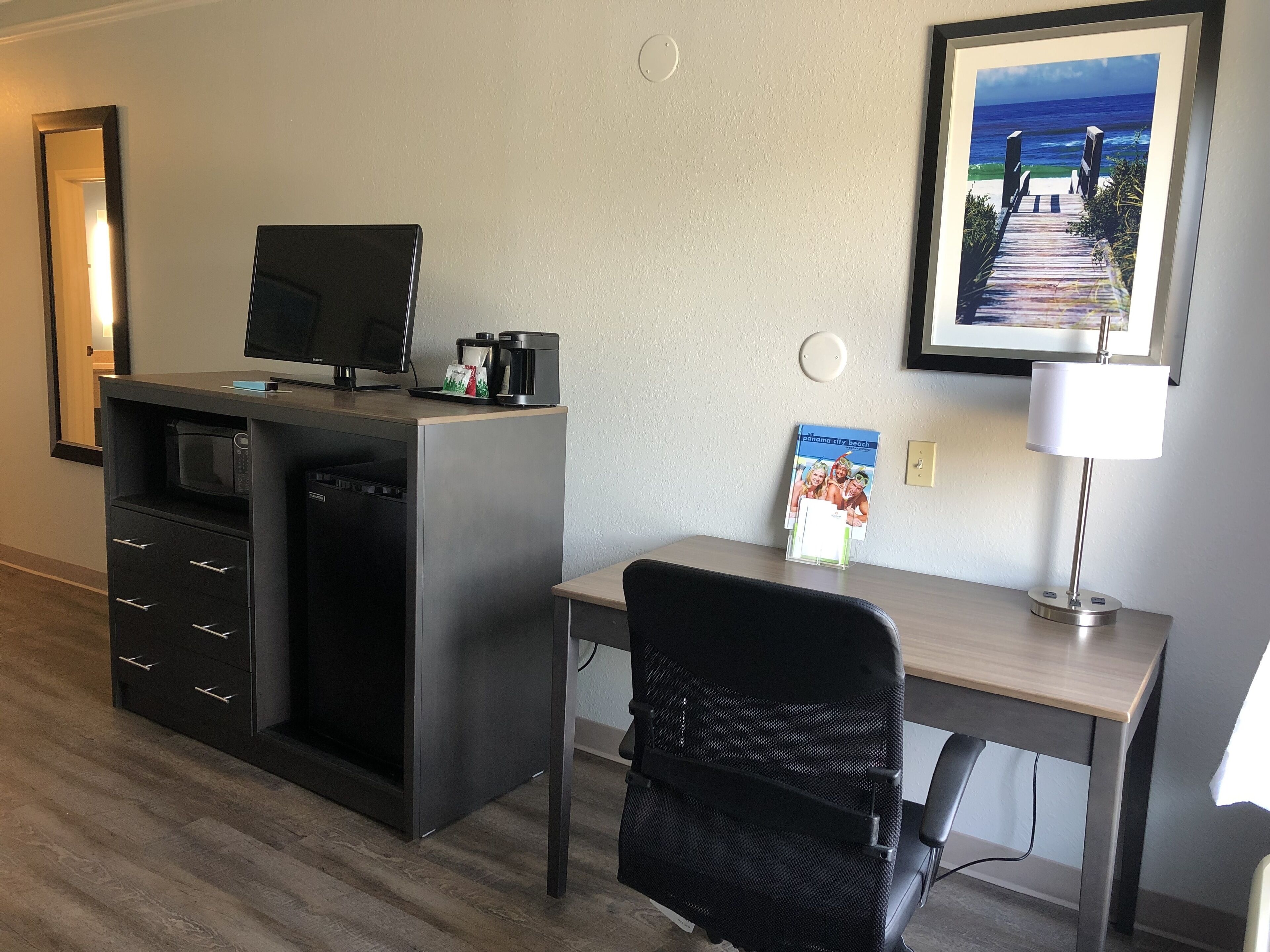 A hotel room with a desk, a chair, a microwave, a coffee maker, and a framed picture of a beach.