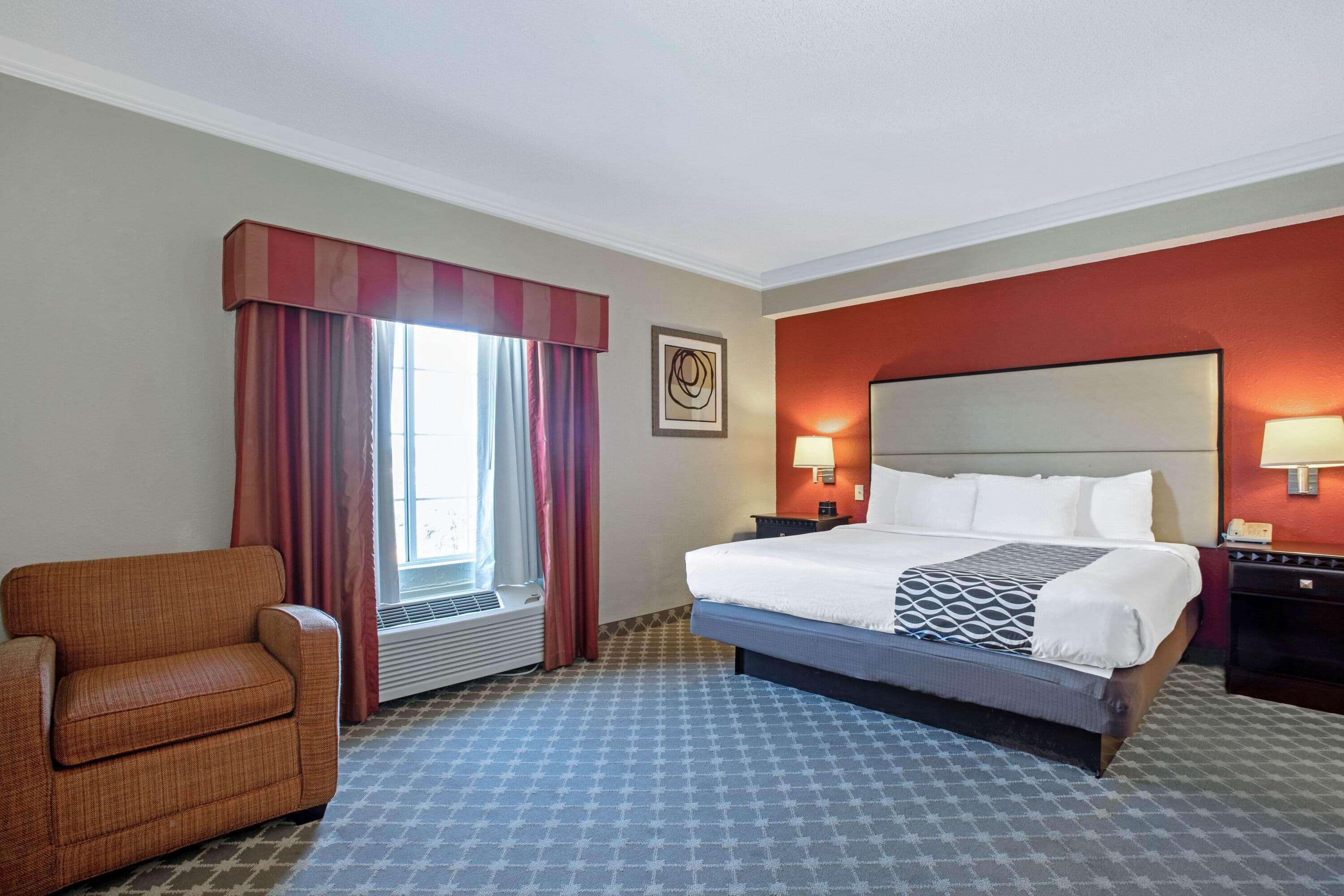 A hotel room with a bed, a chair, a window with curtains, a wall-mounted lamp, and a radiator.
