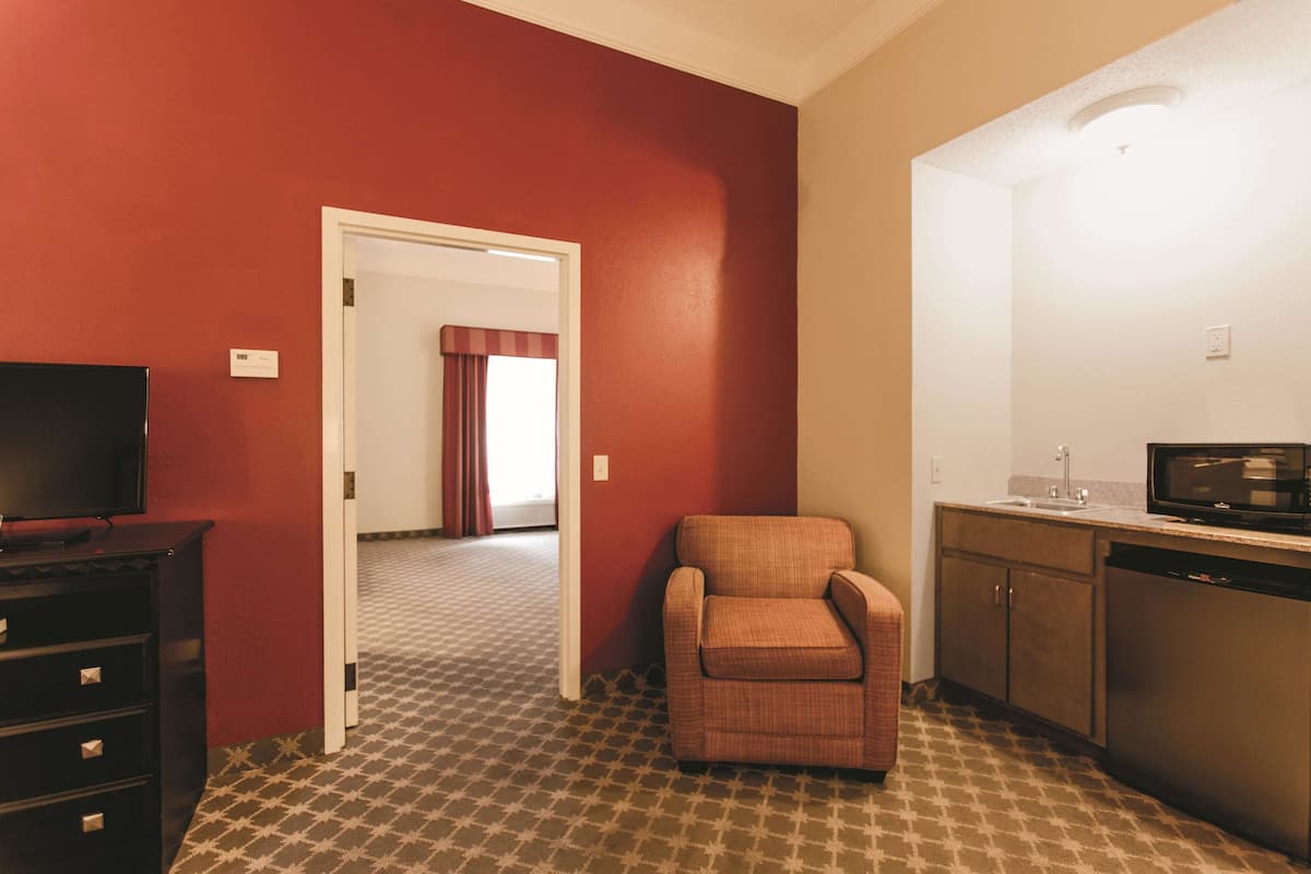 A compact hotel room with a television, a small armchair, a kitchenette, and a patterned carpet.