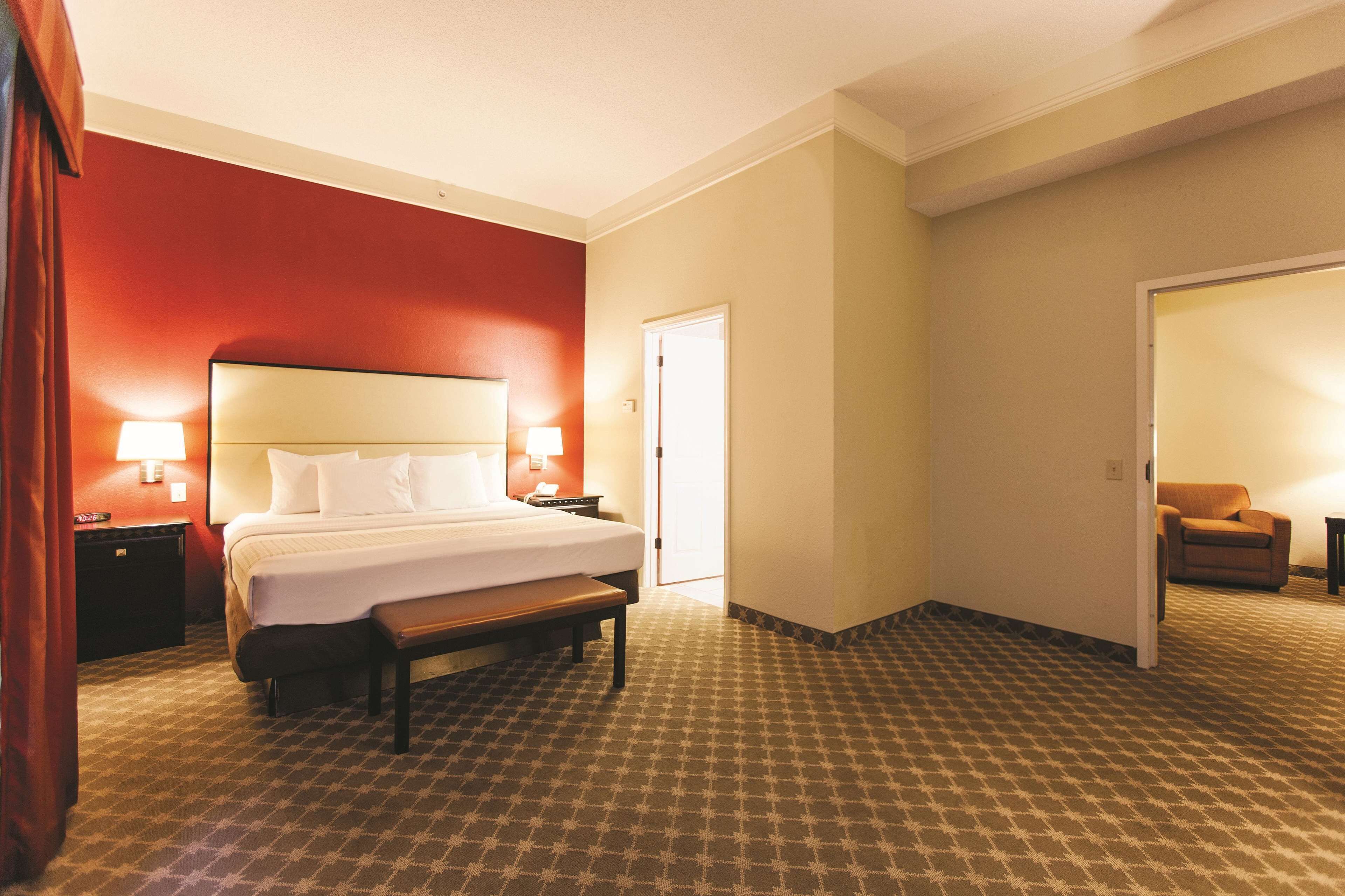 A hotel room with a large bed, bedside lamps, a bench, and a door leading to another room.