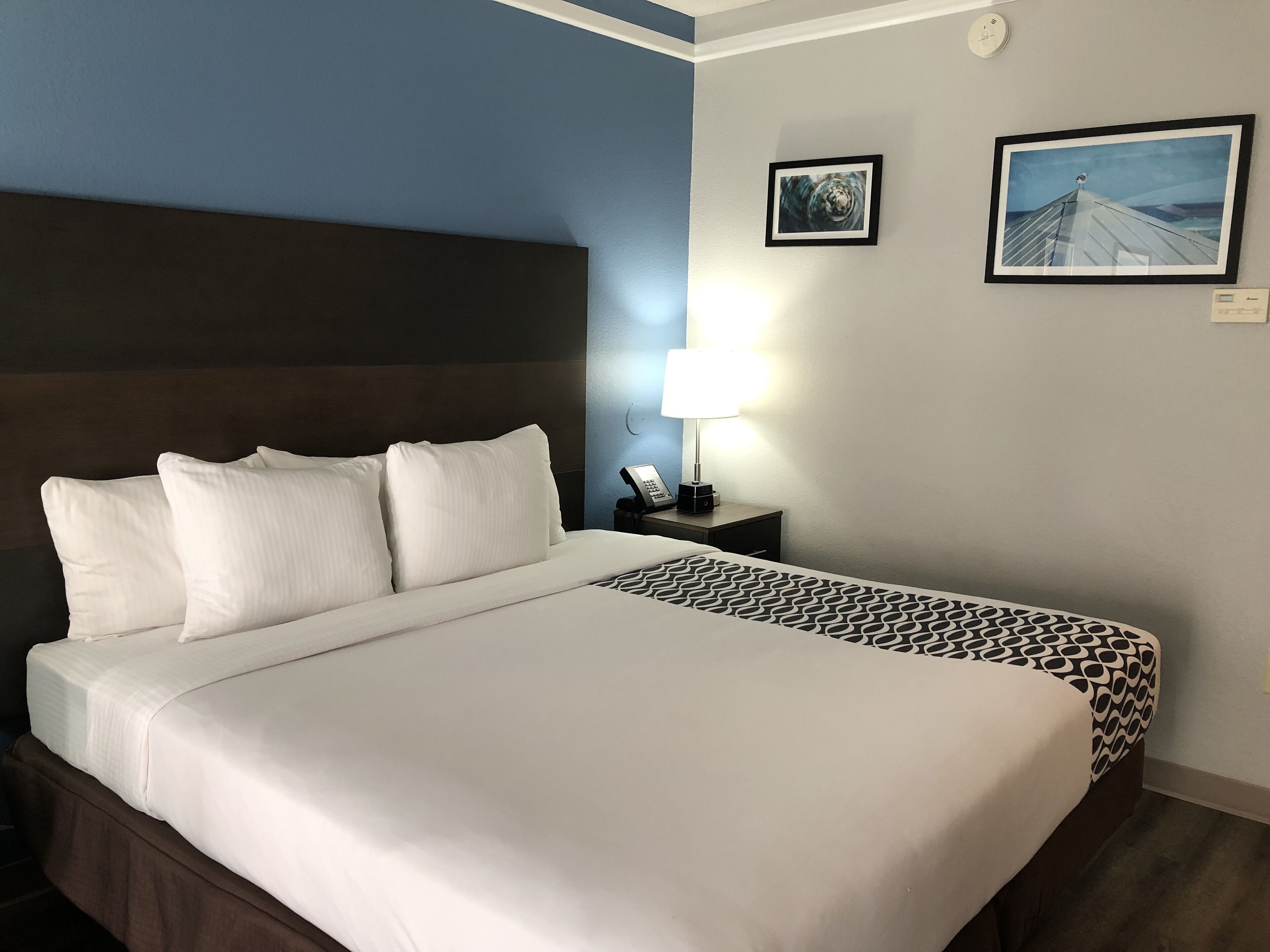 A hotel room with a large bed, a nightstand with a lamp, two framed pictures on the wall, and a clock.
