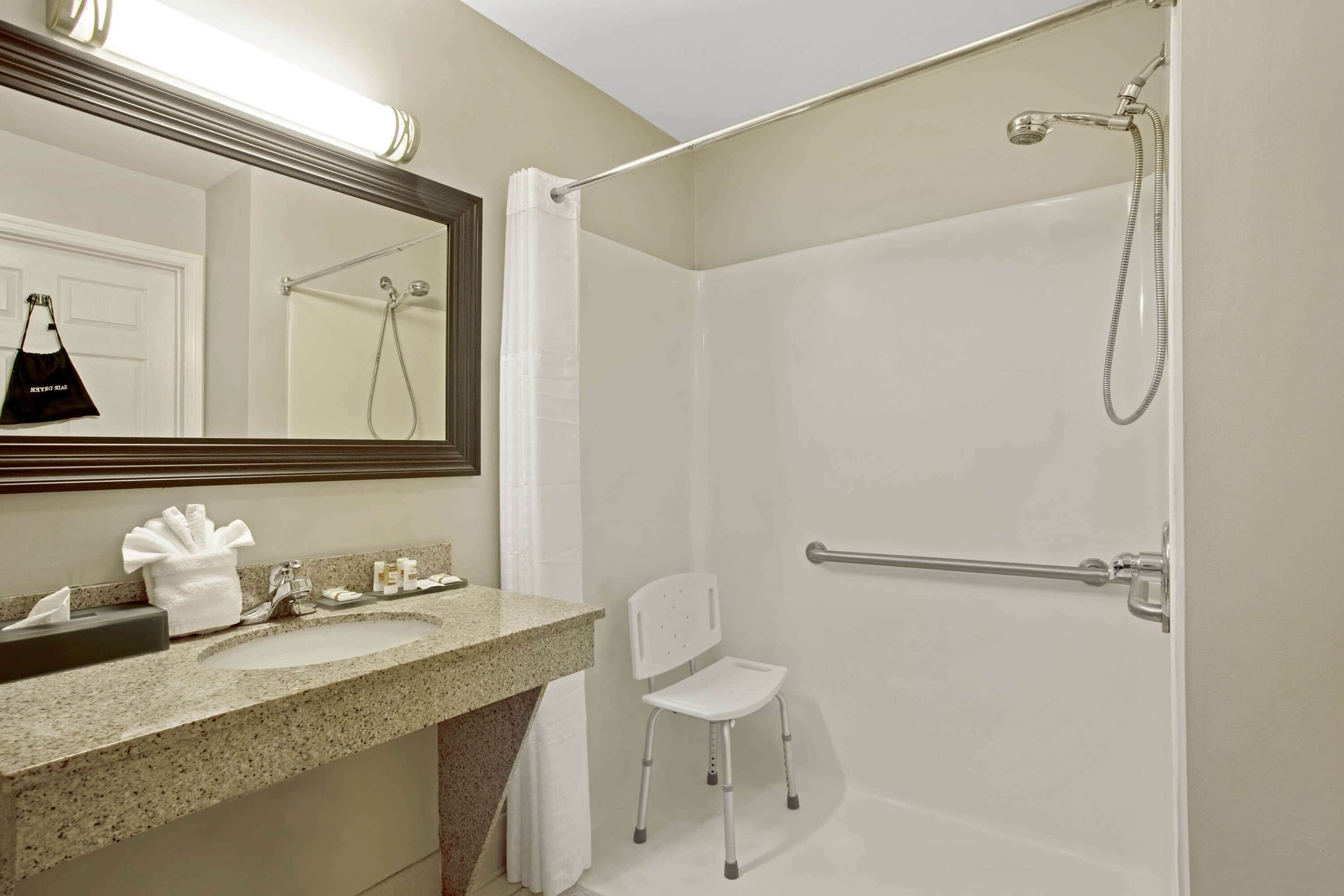 A bathroom with a sink, mirror, shower, and a white chair.