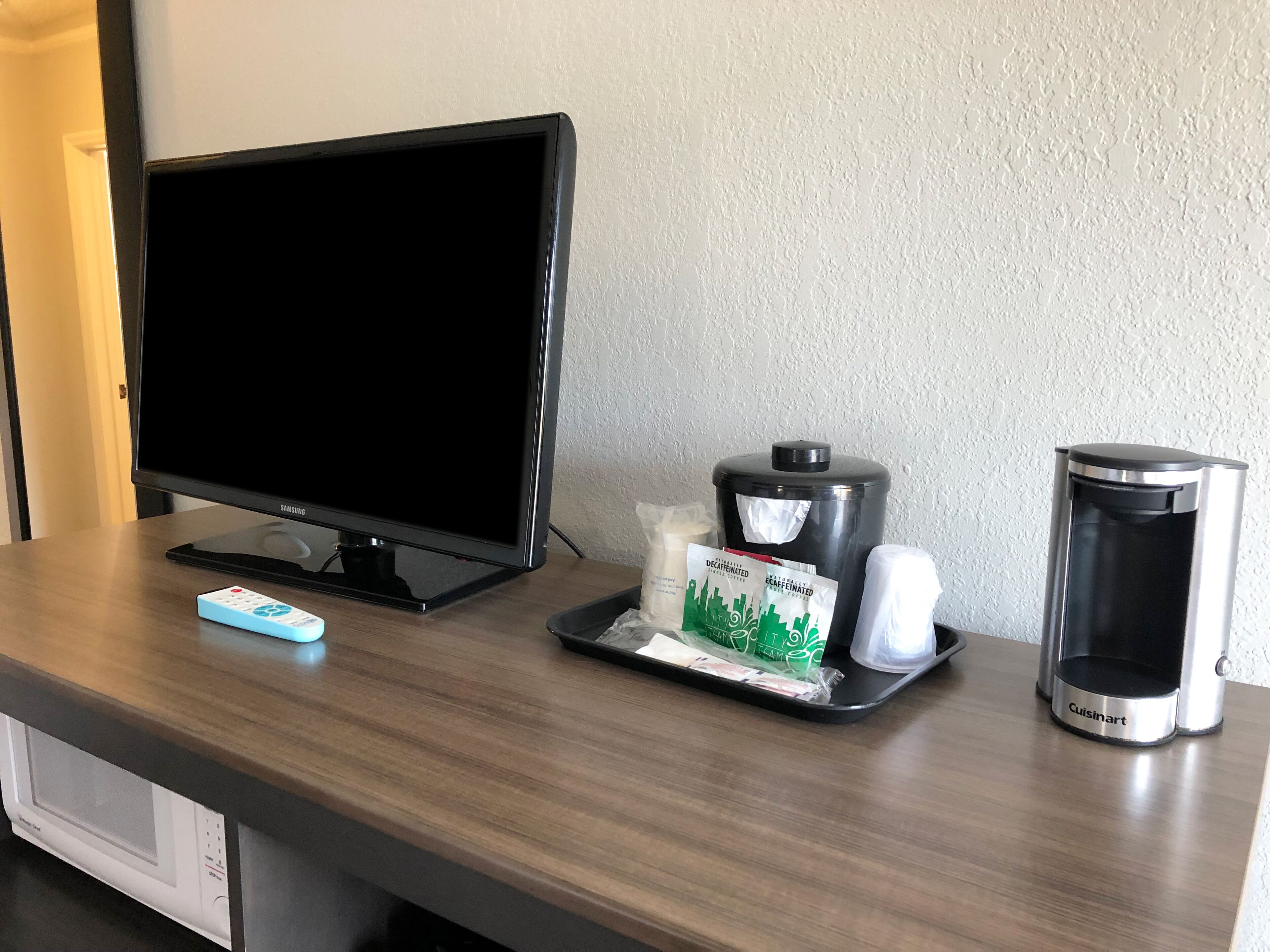 Een bureau in een hotelkamer met een Samsung monitor, een afstandsbediening, een koffiezetapparaat en een prullenbak.