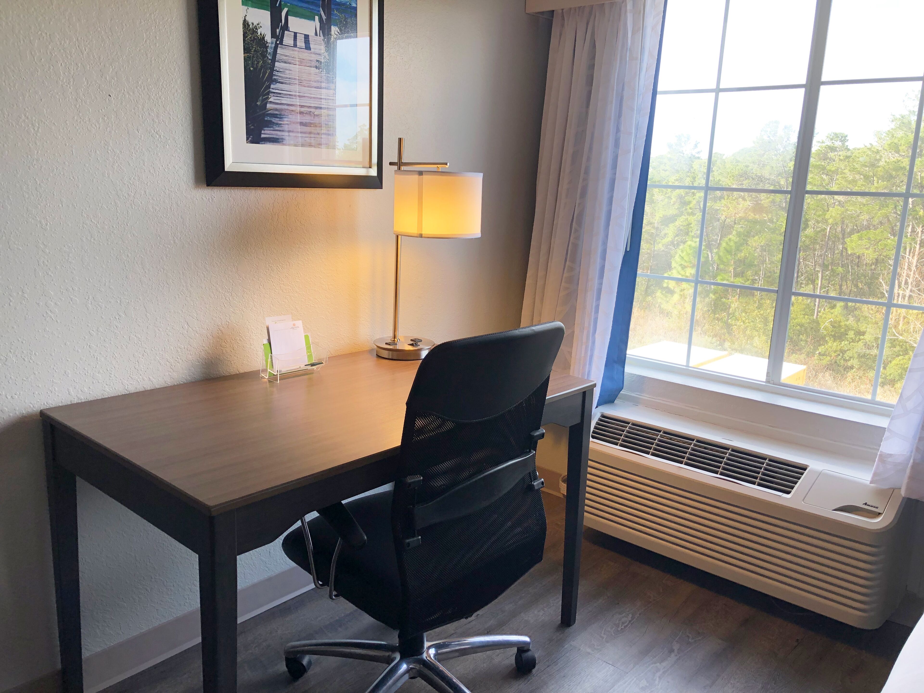 Hotelkamer met een houten bureau, een zwarte bureaustoel, een staande lamp, een raam met uitzicht op bomen en een airconditioner.