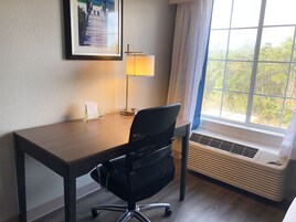 A hotel room with a wooden desk, a black office chair, a standing lamp, a window with a view of trees, and an air conditioning unit.