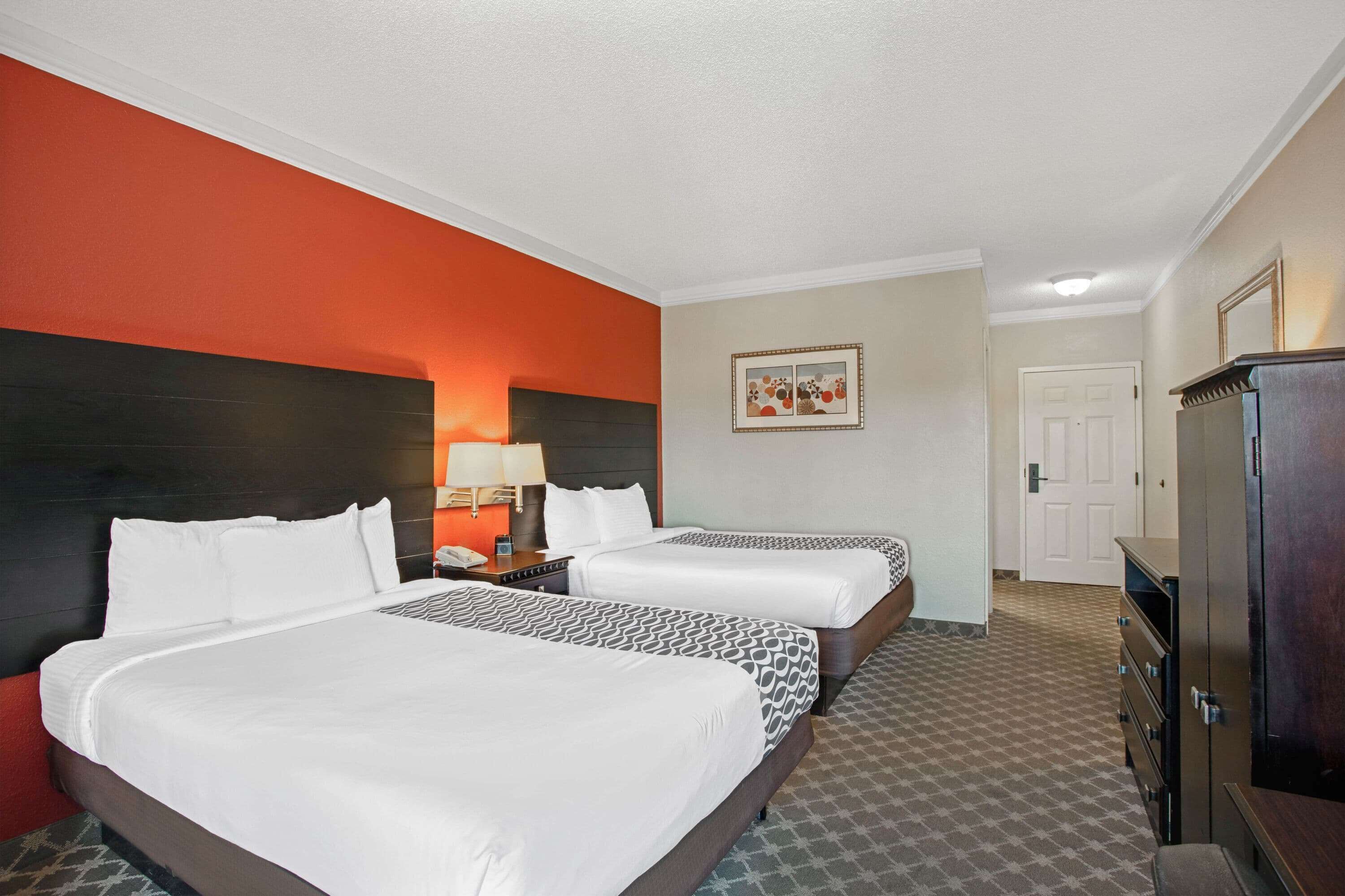A hotel room with two beds, a nightstand, a lamp, a dresser, and a door.