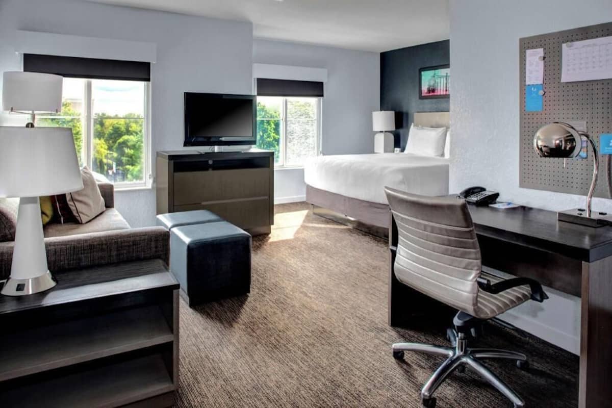 A hotel room with a bed, desk, chair, TV, and a wall-mounted bulletin board.