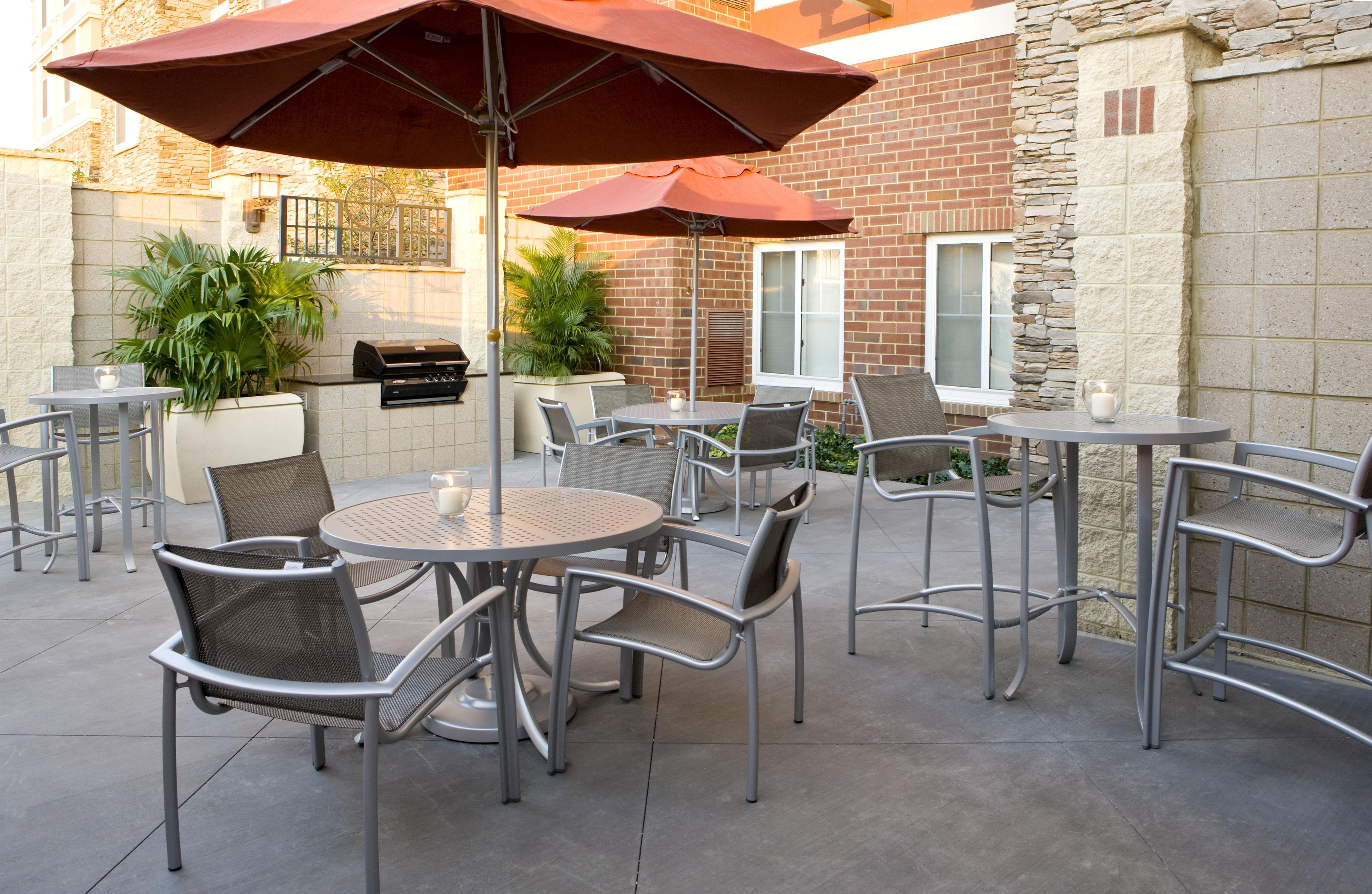 An outdoor patio area with round tables and chairs, a barbecue grill, and umbrellas providing shade.