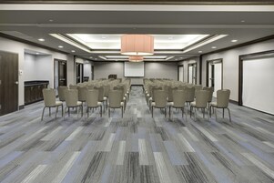Un salón de conferencias con filas de sillas, una pantalla grande y una lámpara en el techo.