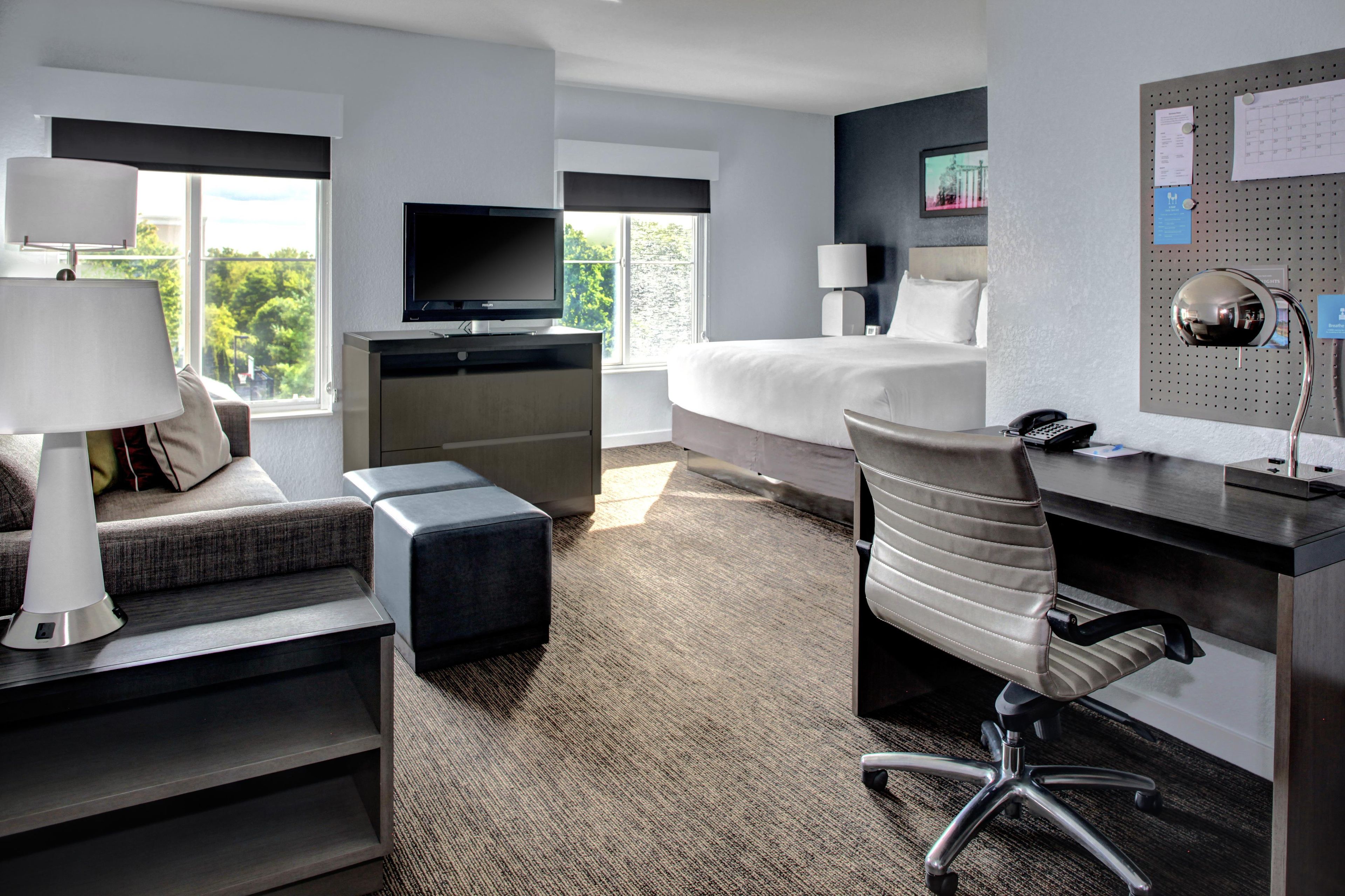A hotel room with a bed, desk, chair, TV, and a view of greenery outside.