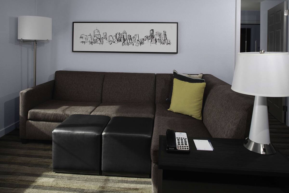 A modern living room with a brown sofa, a black ottoman, a white lamp, and a framed cityscape artwork on the wall.