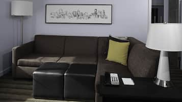 Uma sala de estar moderna com um sofá modular, um pequeno banquinho, uma mesinha de cabeceira com abajur, um controle remoto e uma obra de arte com vista da cidade emoldurada na parede.