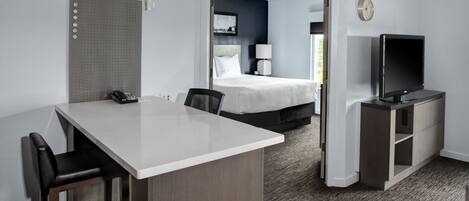 A hotel room with a desk, chairs, a bed, a TV, and a door leading to another room.
