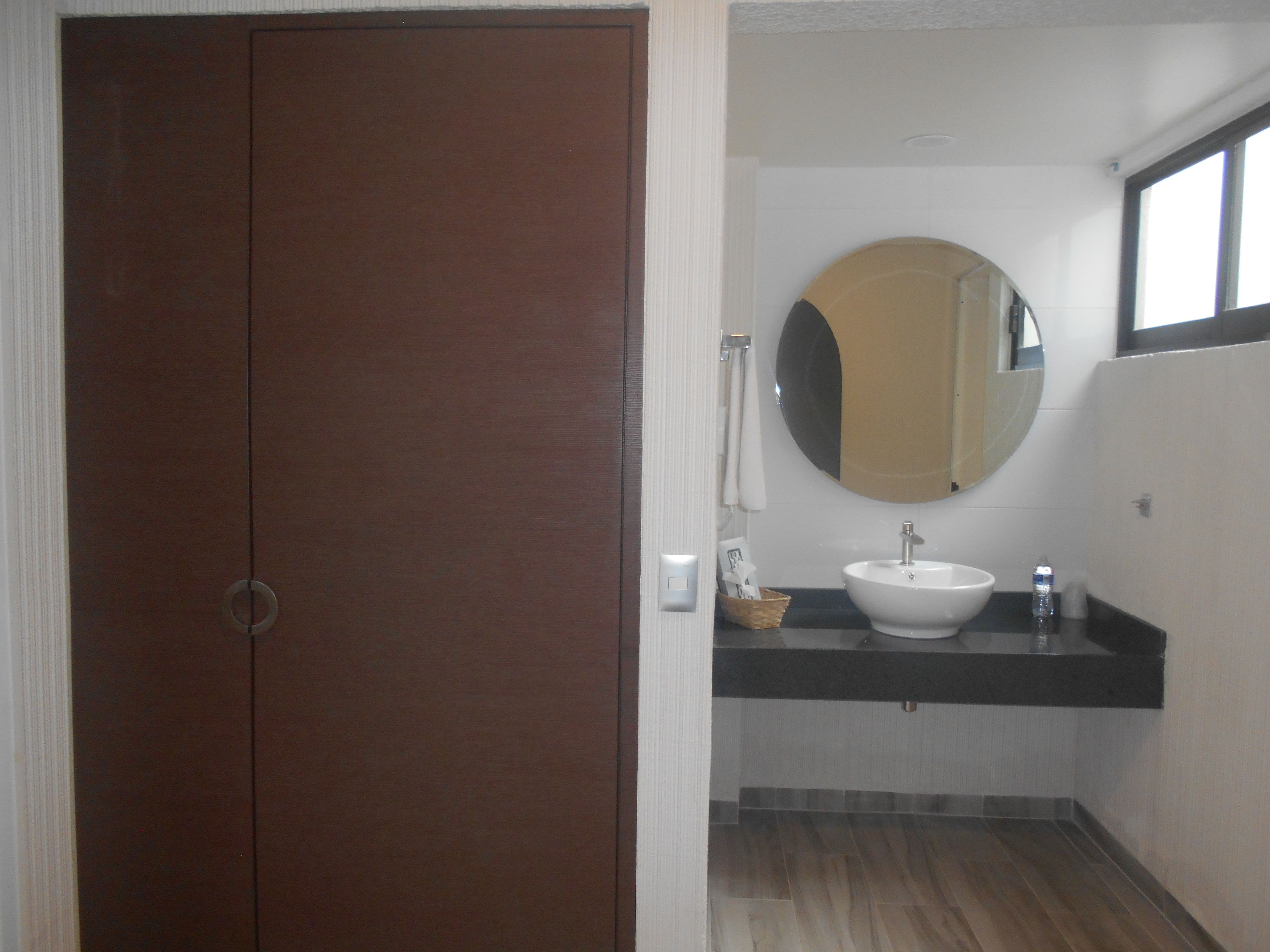 A bathroom with a wooden door, a round mirror, a sink, and a window.