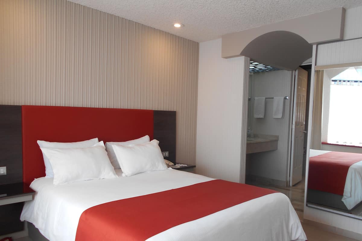 A hotel room with a large bed, a red headboard, a mirror, and a bathroom visible through an open door.