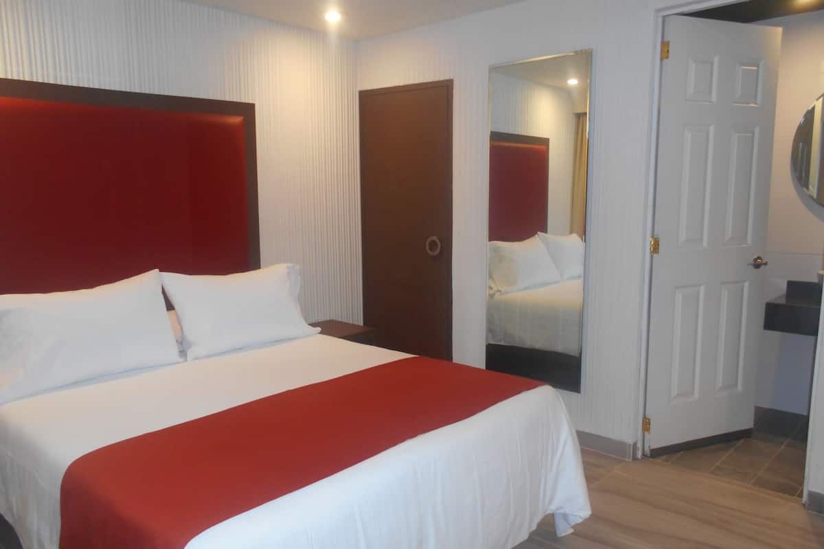 A hotel room with a large bed, a red accent wall, a wooden door, and a mirror.