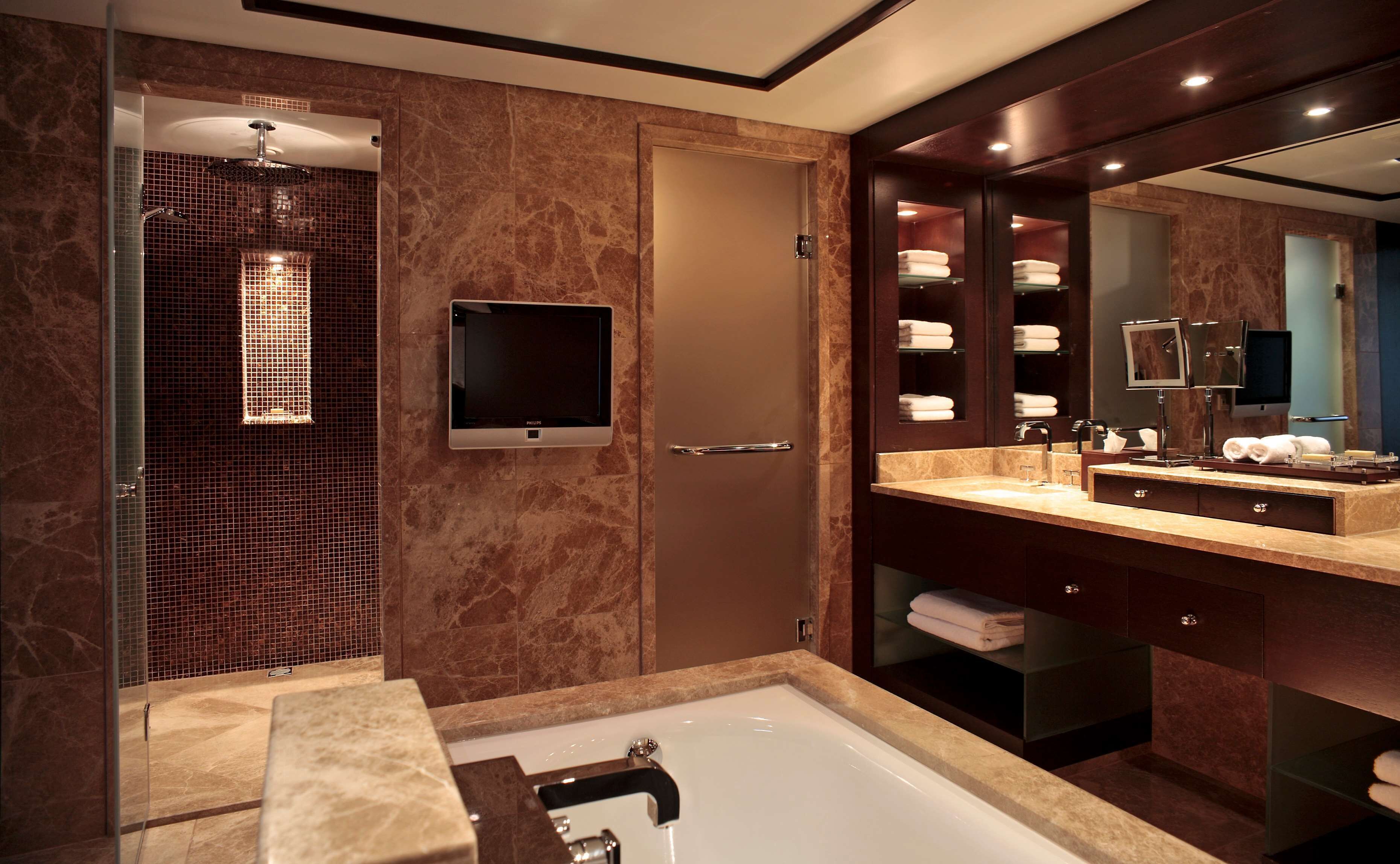 A modern bathroom with a glass-enclosed shower, a large mirror, a flat-screen TV, and a vanity with a sink and towel rack.