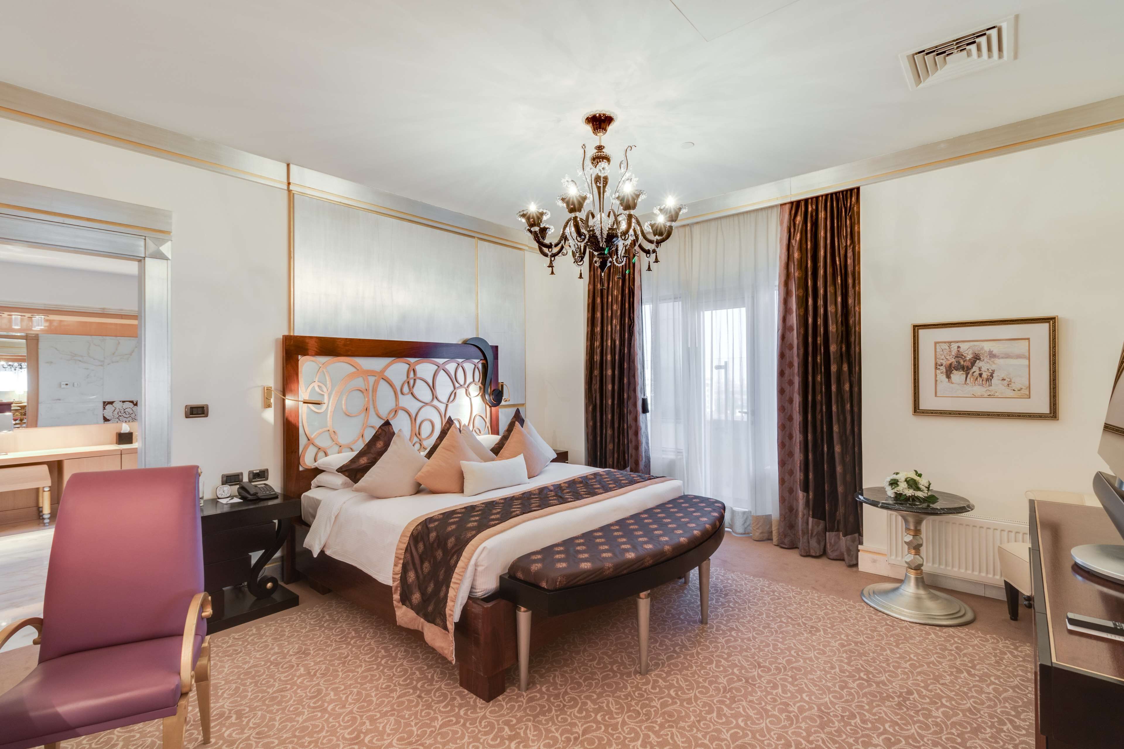 A hotel room with a large bed, a desk, a chair, a chandelier, and a window with curtains.