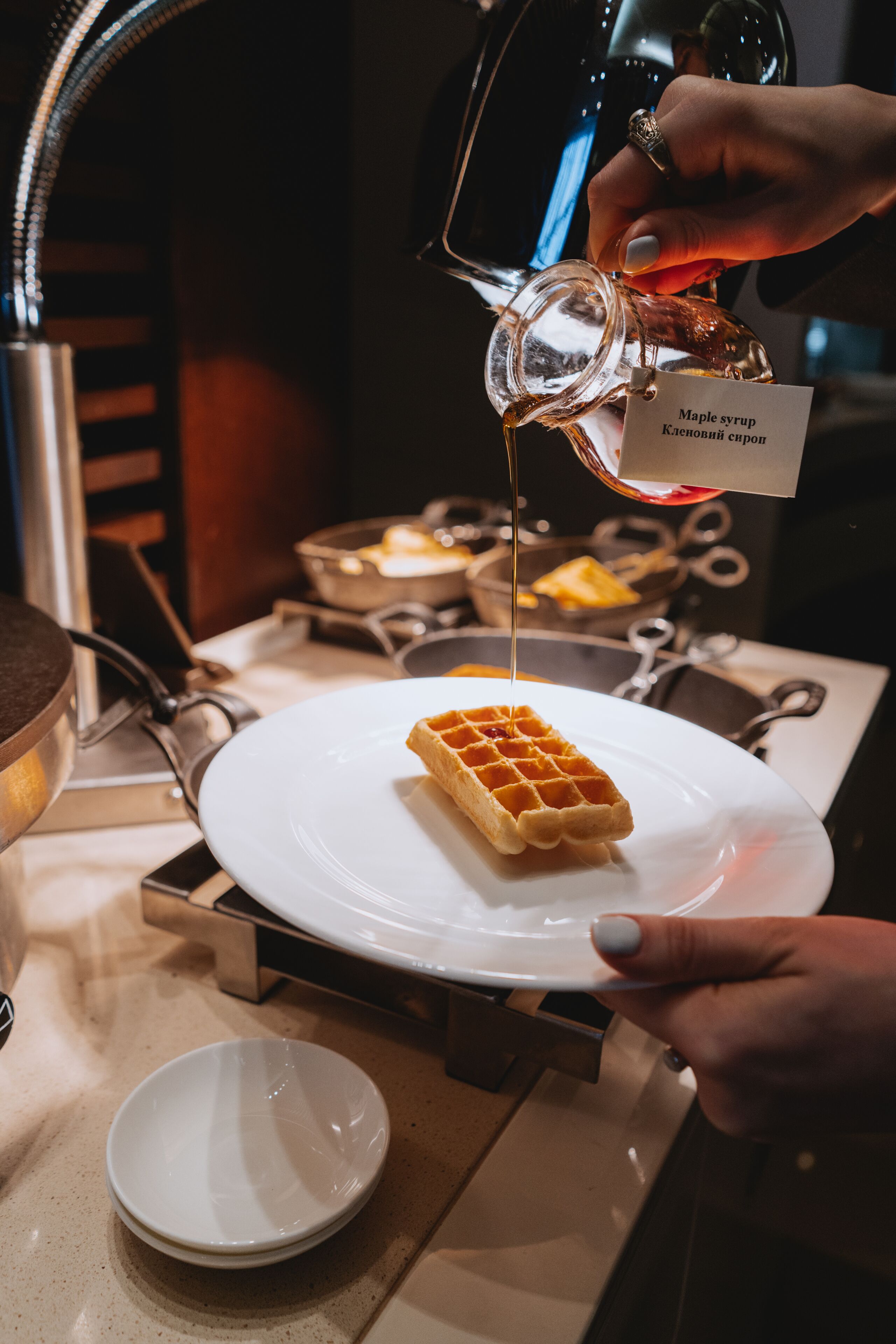 A person pouring maple syrup onto a waffle on a white plate.