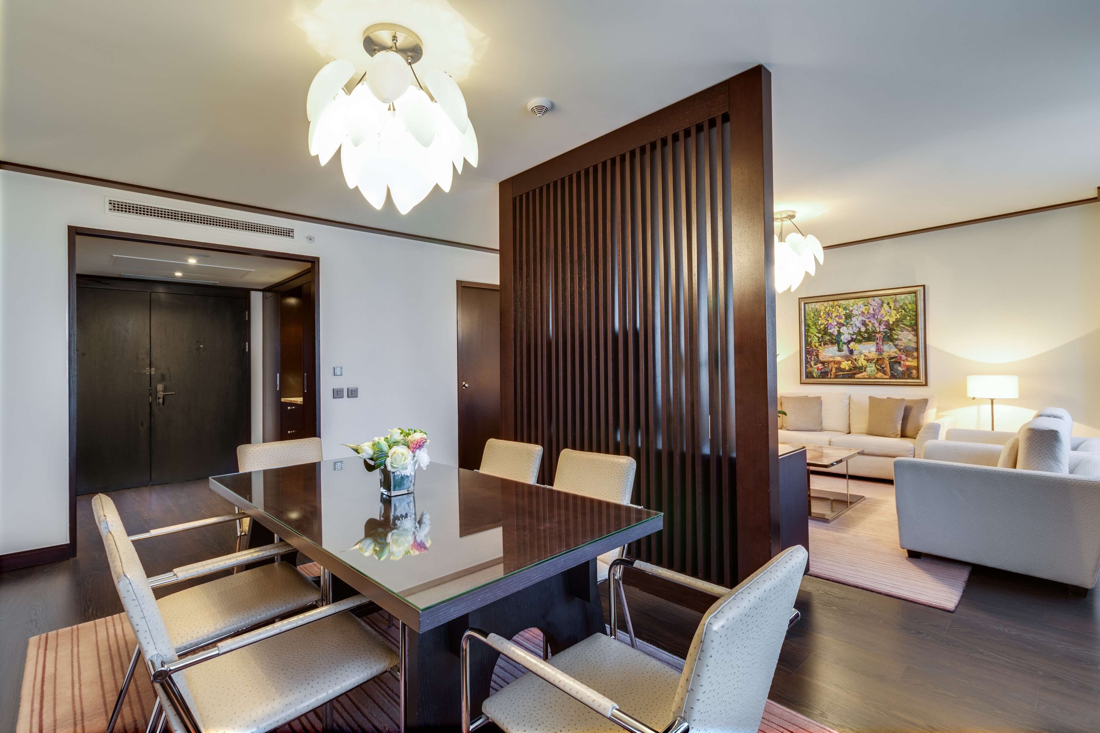 Una habitación de hotel moderna con una mesa de comedor de vidrio, asientos cómodos, una pared de acento de madera y un rincón acogedor con un sofá y una lámpara.