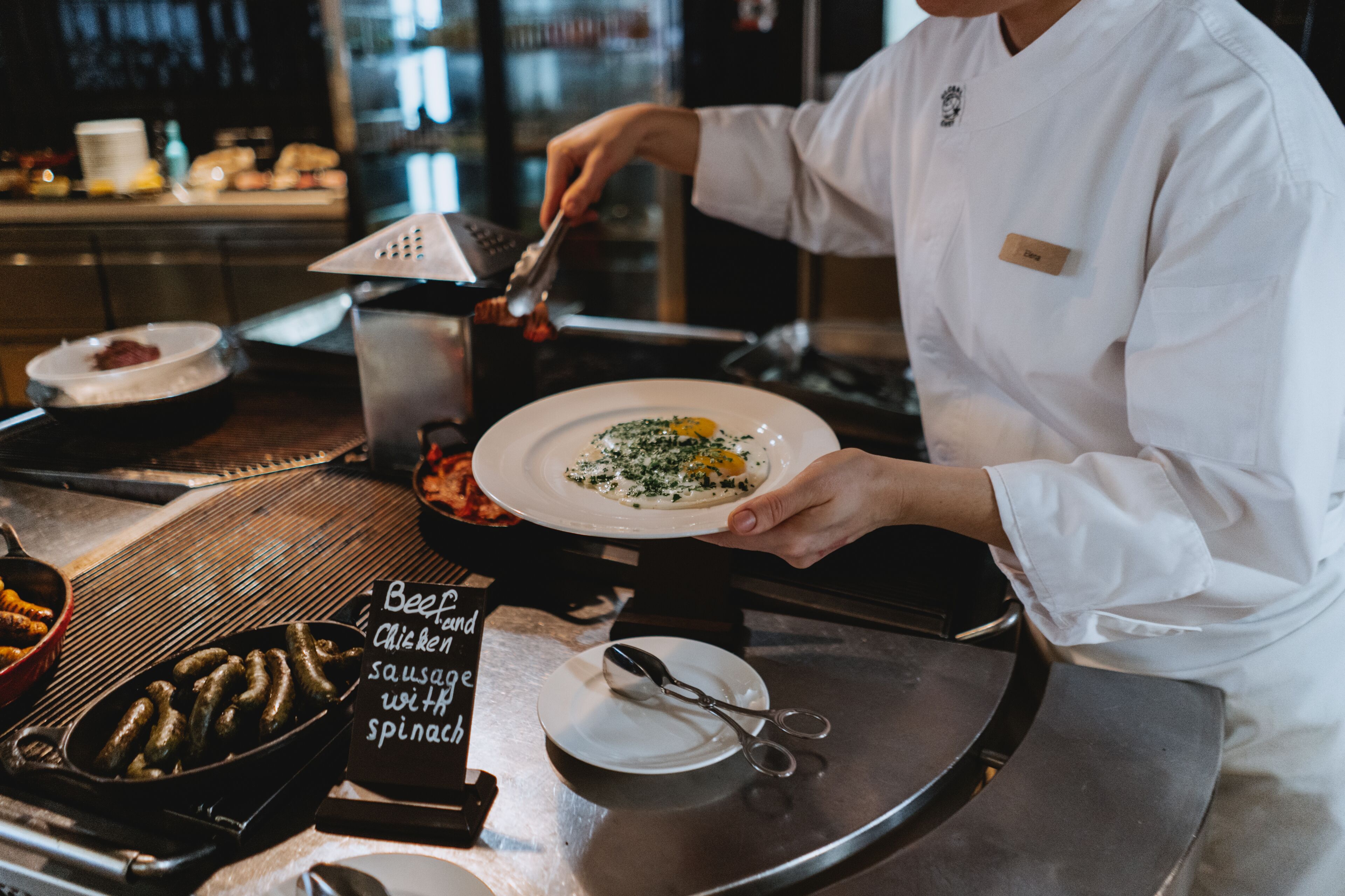 A chef preparing a dish on a stainless steel counter with a name tag, a sign indicating beef, chicken, and sausage with spinach, and a pan of sausages.