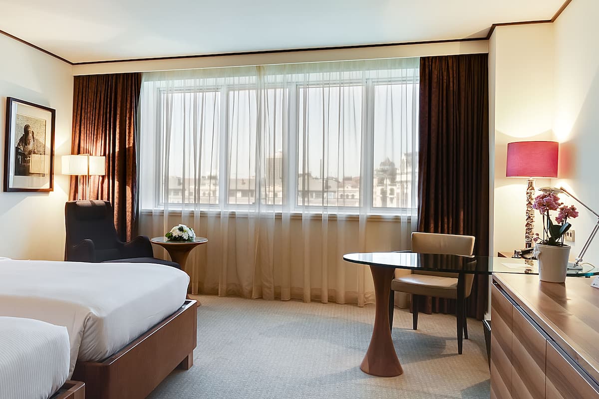 Ein Hotelzimmer mit Bett, Schreibtisch, Stuhl, Lampe und einem Fenster mit Vorhängen.